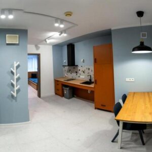 Wnętrze nowoczesnego pokoju w akademiku Uniwersytetu w Siedlcach z dostosowaniem dla osób z niepełnosprawnością ruchową — widoczne biurko, aneks kuchenny i wejście do sypialni.